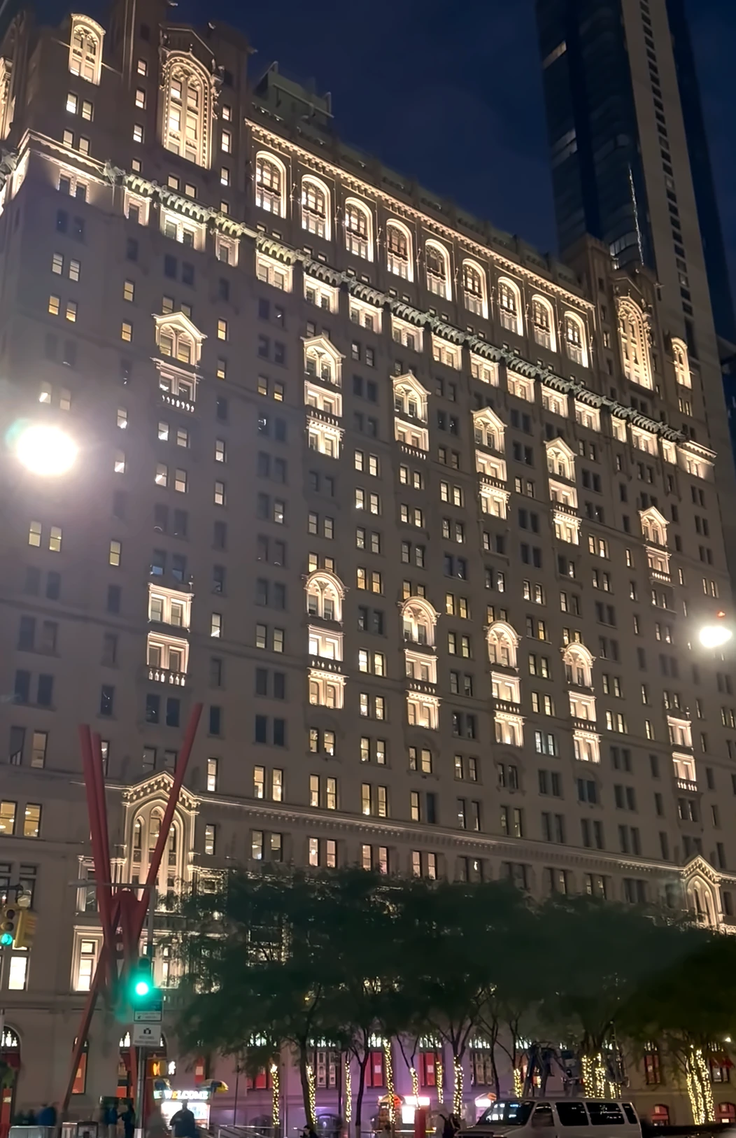 A 21-story pale stone office tower with arched windows lit up at night, trees and a red steel sculpture at its base and a dark glass tower behind