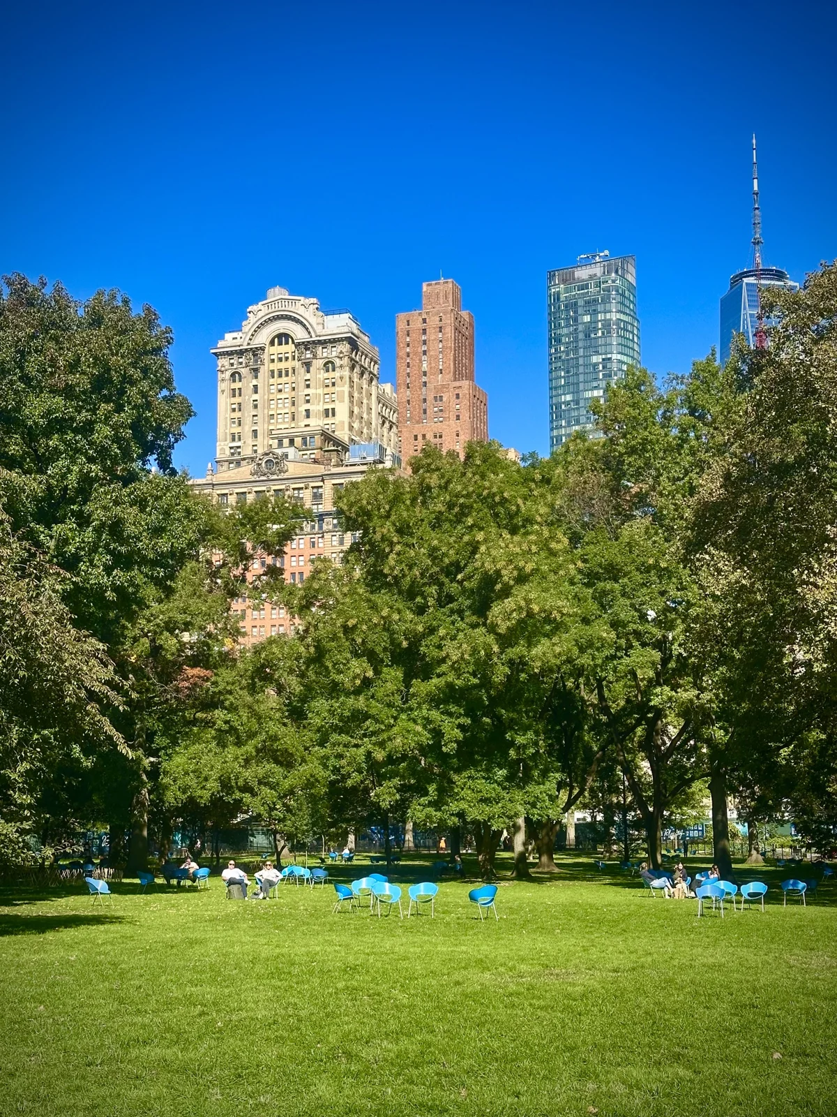 A wide green lawn scattered with bright blue chairs under tall shade trees, with a Beaux-Arts tower, a brick Art Deco tower, a glass tower and One World Trade Center rising behind