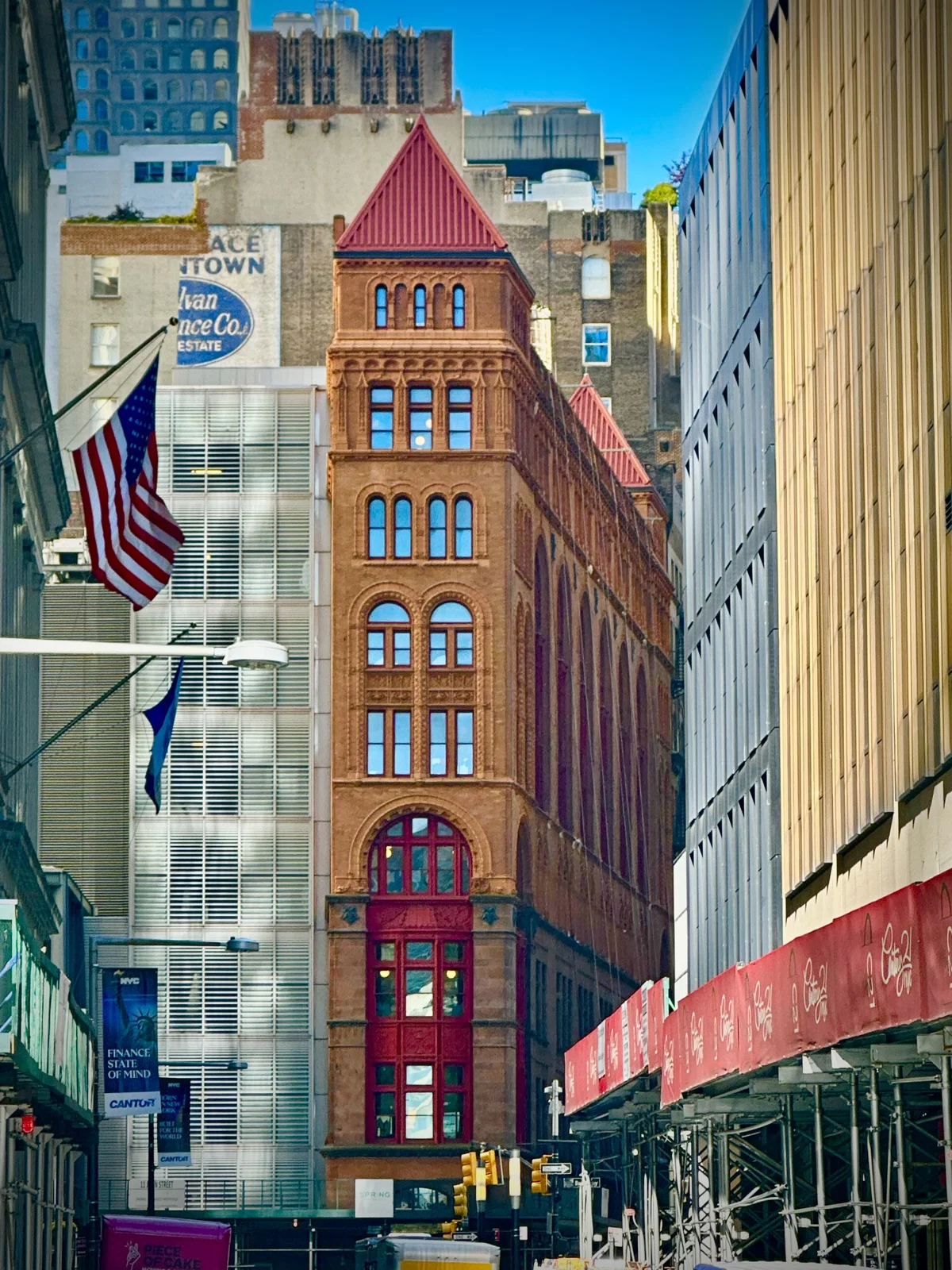 A narrow red-brown terra-cotta and brownstone building with arched windows and a red pyramid roof, between modern towers