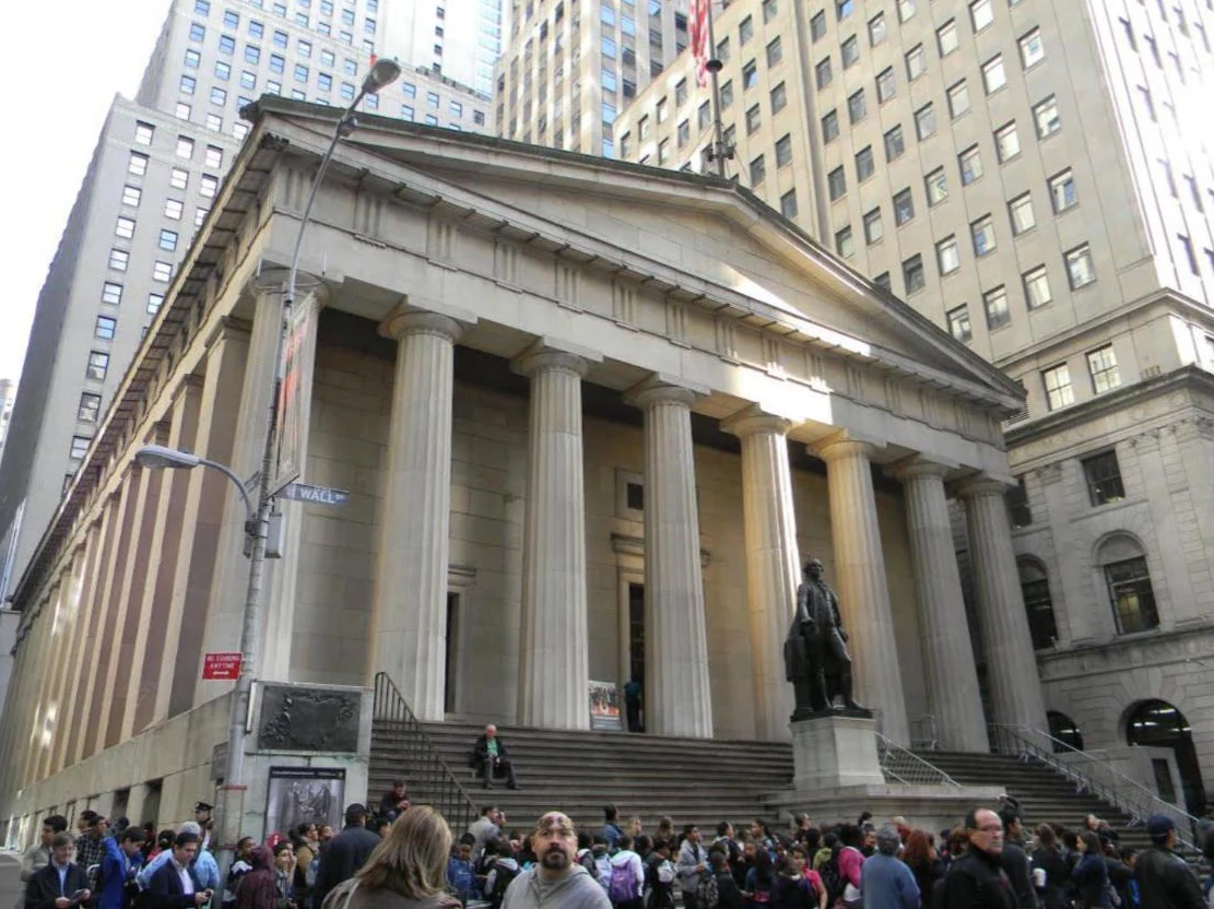 A white marble Greek temple facade with Doric columns and a bronze statue of George Washington, crowds on the steps