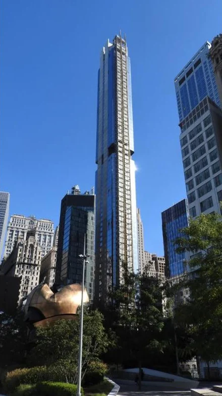 A slender glass tower rising high above older buildings, with a bronze sculpture in the foreground