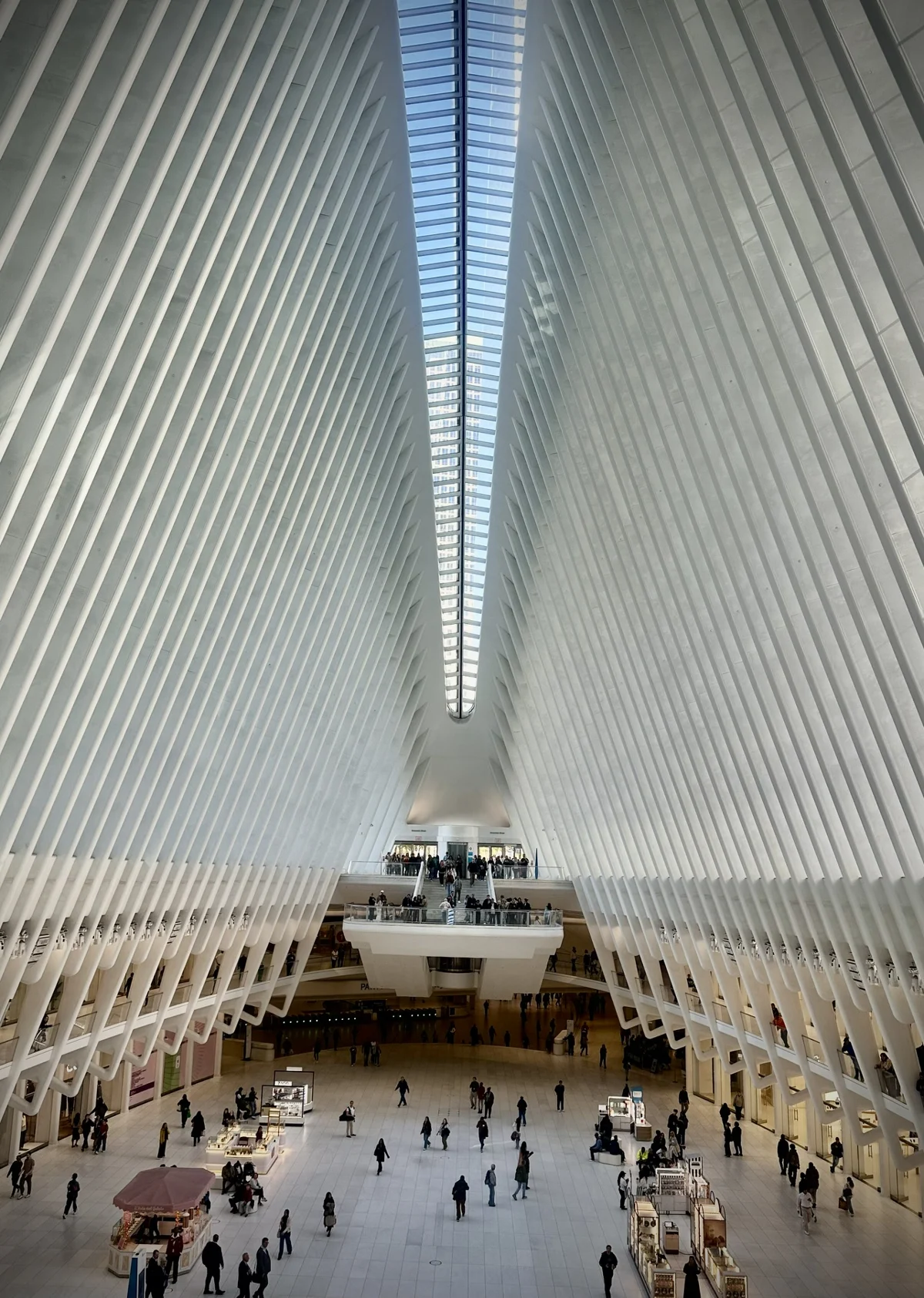 Looking down the vast white ribbed interior of the Oculus toward a long skylight, with people crossing the marble floor far below