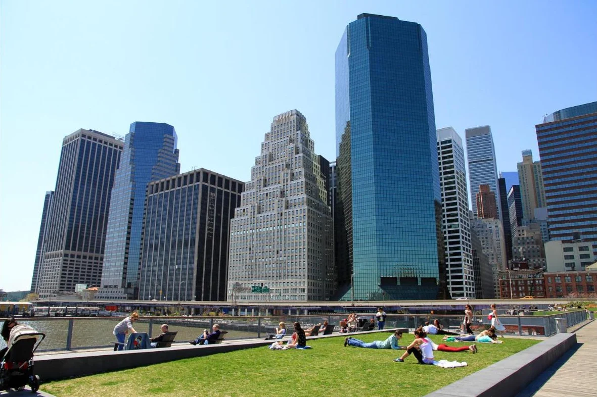 People relaxing on a grassy pier with Financial District towers rising behind them