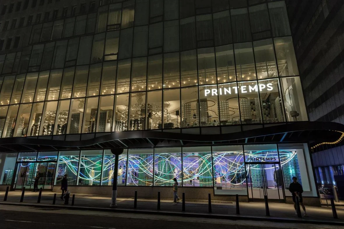 A department store with tall glass windows lit at night, Printemps signs, and neon light art in the windows