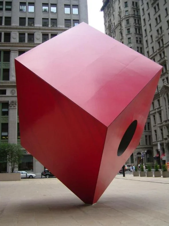 A large red steel cube standing on one point on a plaza, with a round hole through its center