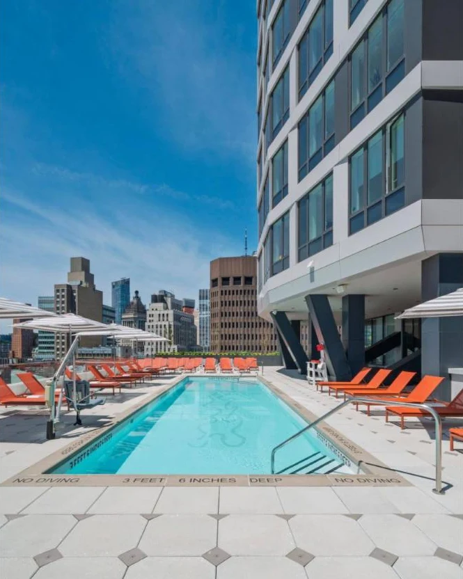 A sunny rooftop pool lined with orange lounge chairs and striped umbrellas, beside the tower