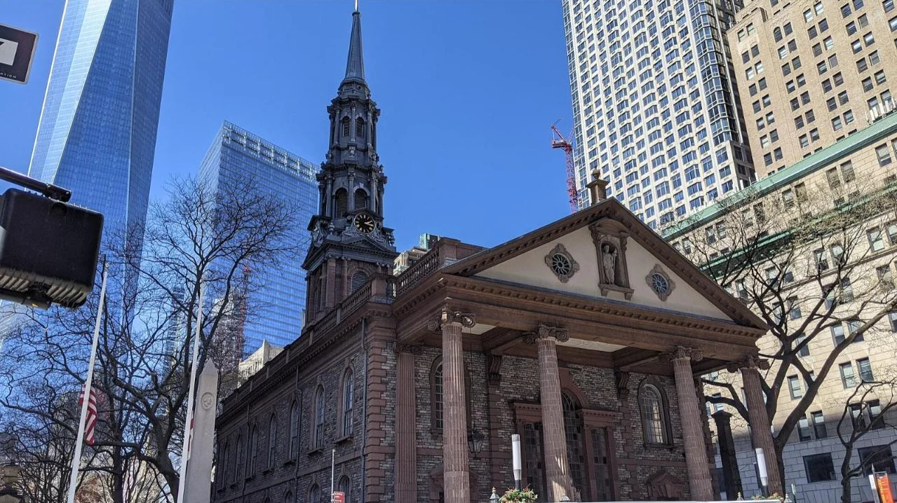A brownstone Georgian chapel with a tall steeple and columned portico, with One World Trade Center rising behind