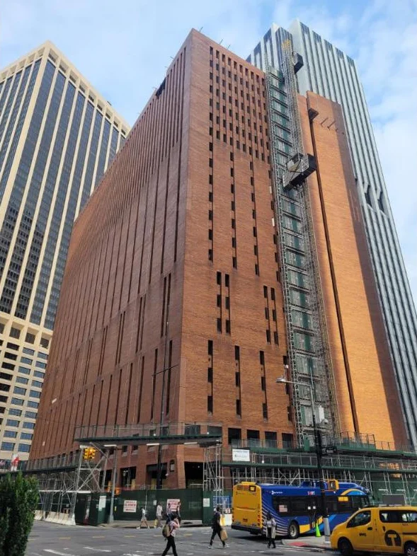 A tall brick office tower wrapped in a construction hoist, with a yellow taxi and a city bus on the street below