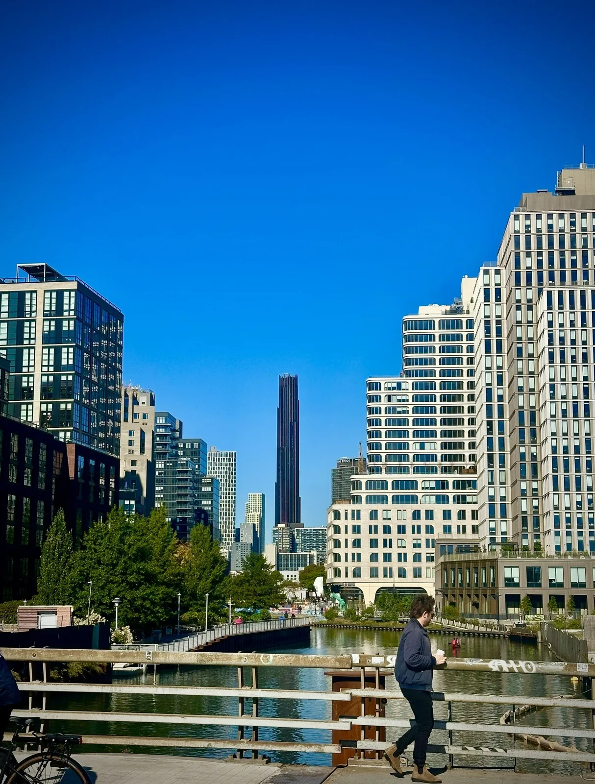 Looking north up the Gowanus Canal from a bridge, new apartment towers and a tree-lined esplanade on both banks, the slender dark Brooklyn Tower rising in the distance under a deep blue sky, a man with a coffee walking along the bridge railing