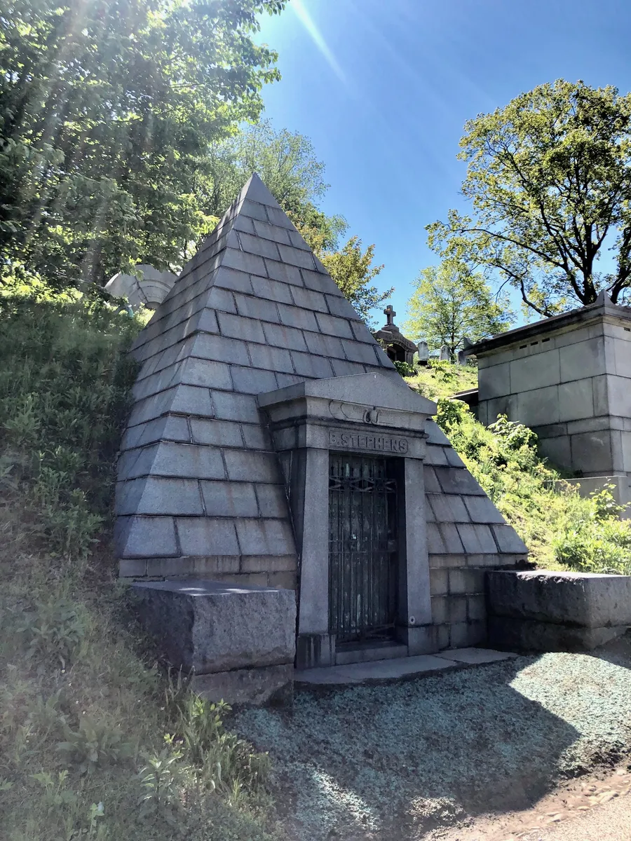 A granite pyramid tomb with a pedimented doorway, set into a wooded hillside beside other mausoleums
